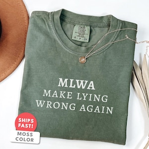 Może przedstawiać: Oliwkowo-zielona koszulka z białym napisem "MLWA MAKE LYING WRONG AGAIN". Złoty naszyjnik z małą zawieszką spoczywa na koszulce. Koszulka jest złożona, a w tle widoczny jest brązowy kapelusz.