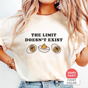 Op de afbeelding: Ivoor kleurig t-shirt met de tekst "THE LIMIT DOESN'T EXIST" in zwarte letters. Onder de tekst staan drie illustraties van gevulde eieren. Het shirt heeft een ronde hals en korte mouwen.