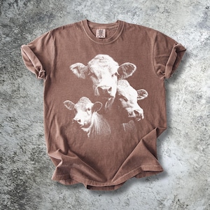 Könnte beinhalten: Braunes T-Shirt mit einer weißen Grafik von drei Kuhgesichtern. Das T-Shirt hat einen Rundhalsausschnitt und kurze Ärmel. Der Stoff hat ein leicht verwaschenes Aussehen.
