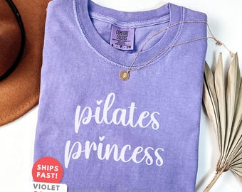 Puede incluir: Una camiseta de color violeta claro con las palabras "pilates princess" en escritura blanca. Un collar dorado con un colgante descansa sobre la camiseta. Un sombrero marrón y una planta seca están en el fondo. La camiseta tiene una etiqueta "Comfort Colors".