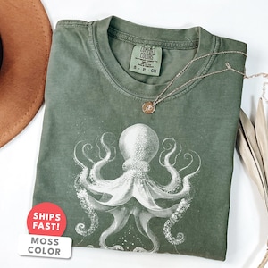 May include: Moss green t-shirt featuring a white octopus design. The shirt has a crew neck and short sleeves. A gold necklace with a small pendant is draped on the shirt. A brown hat and dried palm leaf are in the background.