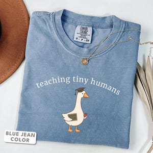 Può includere: Una t-shirt color blu jeans con la grafica di un'oca bianca che indossa un tocco da laurea, mentre tiene una piccola lavagna e una mela rossa. Il testo "teaching tiny humans" è stampato in bianco sopra l'oca. Scollo a girocollo.