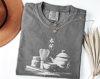 以下が含まれることがあります： グレーのComfort Colors Tシャツ。ティーポット、泡立て器、ボウルなど、お茶の儀式のグラフィックが描かれています。シャツには日本語の文字も含まれています。ペンダント付きのゴールドネックレスがシャツの上に置かれています。