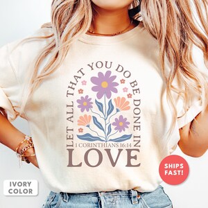 May include: Ivory-colored t-shirt featuring a floral design with the text "LET ALL THAT YOU DO BE DONE IN LOVE" and the Bible verse "1 Corinthians 16:14." The design includes purple and peach flowers with blue leaves.