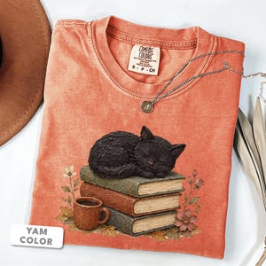 以下が含まれることがあります： コーラルカラーの半袖Tシャツ。3冊の本の山の上で眠る黒猫と、茶色のマグカップ、花のグラフィックがプリントされています。首元にはComfort Colorsのブランドラベルがあり、シルバーのネックレスが添えられています。