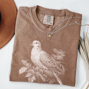 Könnte beinhalten: Ein braunes Comfort Colors T-Shirt mit einer Vogelillustration. Das T-Shirt hat einen Rundhalsausschnitt und kurze Ärmel. Eine silberne Halskette mit einem kleinen Anhänger liegt auf dem T-Shirt. Ein brauner Hut ist im Hintergrund zu sehen.