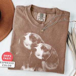 Könnte beinhalten: Espressofarbenes Comfort Colors T-Shirt mit einer Grafik von zwei Hunden. Das Shirt hat einen Rundhalsausschnitt und kurze Ärmel. Eine silberne Halskette mit einem kleinen Anhänger liegt auf dem Shirt. Das Shirt ist auf einer weißen Oberfläche gefaltet.
