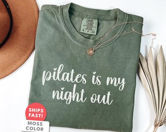 以下が含まれることがあります： モスグリーンのTシャツに、白い筆記体で「pilates is my night out」と書かれています。小さなペンダントが付いたゴールドのネックレスがシャツの上にあります。茶色の帽子と乾燥したヤシの葉も写っています。