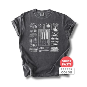 Può includere: T-shirt grigio scuro con un'illustrazione in bianco e nero di utensili e ingredienti da cucina. Il design include coltelli, un tagliere, verdure, spezie e cucchiai. La maglietta ha le maniche arrotolate e un'etichetta con "Pepper Color".