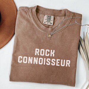 Op de afbeelding: Een bruin t-shirt met de woorden "ROCK CONNOISSEUR" in het wit gedrukt. Een zilveren ketting met een kleine hanger ligt op het shirt. Een bruine hoed en gedroogde palmbladeren zijn op de achtergrond.