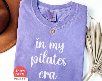 Può includere: Una maglietta viola con la scritta "in my pilates era" in corsivo bianco. Una collana d'oro con un ciondolo è appoggiata sulla maglietta. La maglietta ha un girocollo e maniche corte. Un cappello marrone e una pianta secca sono sullo sfondo.
