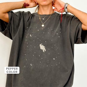Könnte beinhalten: Ein pfefferfarbenes T-Shirt mit einem Grafikdruck eines Astronauten, der im Weltraum schwebt, umgeben von Sternen, einer Mondsichel und einem kleinen Planeten. Das Design ist mittig auf der Brust des Kleidungsstücks mit Rundhalsausschnitt.