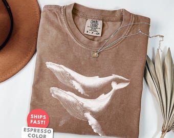 May include: Espresso-colored t-shirt featuring a detailed white illustration of two whales. The shirt has a crew neck and short sleeves. A silver necklace with a small pendant rests on the shirt. The tag reads "Comfort Colors".