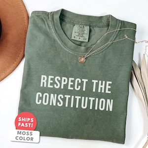 Puede incluir: Una camiseta verde musgo con el texto blanco "RESPECT THE CONSTITUTION". Un collar dorado con un colgante descansa sobre la camiseta. Un sombrero marrón y hojas de palma secas están en el fondo.