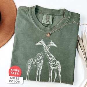 May include: A moss green t-shirt featuring a graphic of three giraffes. The shirt has a crew neck and short sleeves. A gold necklace with a pendant is draped across the shirt. The shirt is labeled "Comfort Colors".