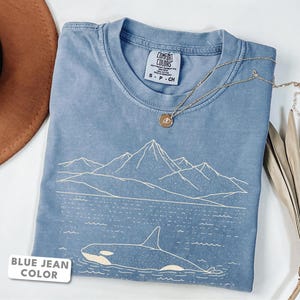 Könnte beinhalten: Ein blaues T-Shirt mit einer weißen Linienzeichnung eines Orcas, der im Wasser vor einer Bergkulisse schwimmt. Das T-Shirt hat einen Rundhalsausschnitt und ein Comfort Colors Markenetikett am Innenkragen mit der aufgedruckten Größe S-CH.