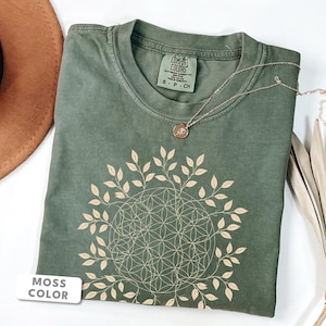 Könnte beinhalten: Ein moosgrünes Kurzarm-T-Shirt mit einer cremefarbenen Grafik eines geometrischen Lebensblumenmusters, das von einem kreisförmigen Blattkranz umgeben ist. Das T-Shirt hat einen Rundhalsausschnitt und ein Etikett mit der Aufschrift Comfort Colors S-P-CH.