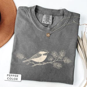 May include: A pepper-colored short-sleeve t-shirt featuring a light-colored line art illustration of a chickadee perched on a pine branch. The shirt has a crew neckline and a Comfort Colors brand label on the inside collar with size S-P-CH.