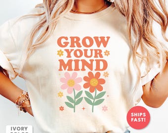 Op de afbeelding: Ivoor kleurig t-shirt met de tekst "GROW YOUR MIND" in oranje. Onder de tekst staan twee bloemen, een roze en een oranje, met groene stelen en bladeren. Het shirt heeft een ronde hals.