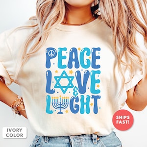 May include: Ivory-colored t-shirt with a blue and turquoise graphic design. The design includes the words "PEACE LOVE & LIGHT" with a peace symbol, a Star of David, and a menorah. The shirt has a round neck and short sleeves.