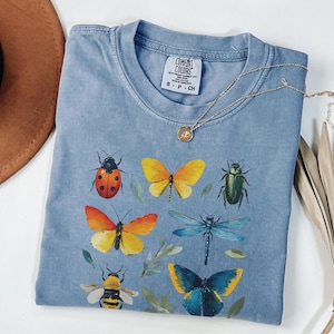 Op de afbeelding: Een lichtblauw Comfort Colors T-shirt met kleurrijke insectenillustraties, waaronder een lieveheersbeestje, vlinders, een kever, een libelle en een bij. Een gouden ketting met een hanger ligt op het shirt. Het shirt ligt op een witte ondergrond.
