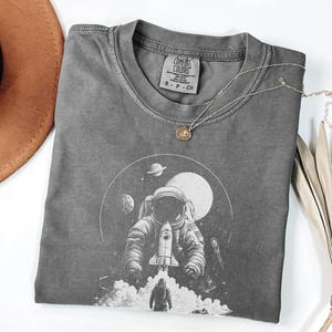 May include: A gray Comfort Colors t-shirt featuring a detailed graphic of an astronaut with a rocket ship, planets, and a moon. A silver necklace with a small pendant rests on the shirt. A brown hat and dried palm leaf are in the background.