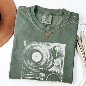 May include: An olive green t-shirt with a graphic of a vintage record player. The shirt has a crew neck and a small tag on the collar. A gold necklace with a small pendant rests on the shirt. A brown hat and dried palm leaves are in the background.