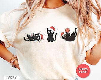 Puede incluir: Camiseta de color marfil con tres ilustraciones de gatos negros con gorros de Papá Noel. Un gato está enredado en luces navideñas, otro está sentado erguido y el tercero juega con un adorno. La camiseta tiene el texto "IVORY COLOR".