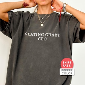 Puede incluir: Camiseta gris oscuro de gran tamaño con el texto "SEATING CHART CEO" en blanco. La camiseta tiene mangas cortas y un corte holgado. Una pegatina roja y blanca que dice "SHIPS FAST!" y "PEPPER COLOR" está en la parte inferior derecha.