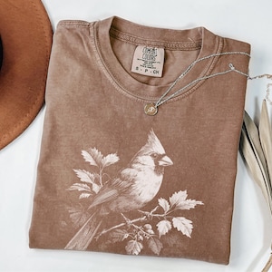 May include: A brown Comfort Colors t-shirt with a bird illustration on the front. A silver necklace with a small pendant is draped across the shirt. A brown hat is visible in the upper left corner. The shirt has a crew neck.