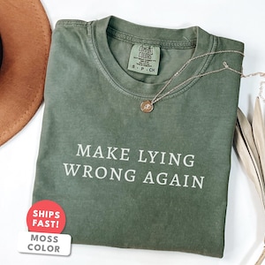 Może przedstawiać: Zielona koszulka w kolorze mchu z białym napisem "MAKE LYING WRONG AGAIN". Złoty naszyjnik z małą zawieszką spoczywa na koszulce. Brązowy kapelusz i suszona roślina znajdują się w tle.