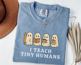 May include: A light blue Comfort Colors t-shirt with the text "I TEACH TINY HUMANS". The shirt features a graphic of four ghost characters holding a pencil, book, coffee mug, and clipboard. A gold necklace is draped across the shirt.
