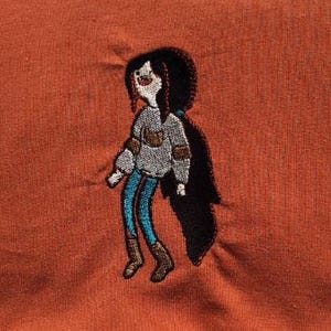 Puede incluir: Diseño bordado de un personaje de dibujos animados sobre tela naranja. El personaje tiene pelo largo negro, un suéter gris con un bolsillo marrón, pantalones azules y botas marrones. El diseño está detallado con detalles blancos y rojos.