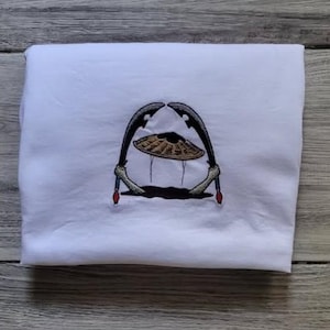 May include: White sweatshirt with an embroidered design. The design features a stylized image of a hat with two curved blades on either side. The blades are black with a gold-coloured hat in the centre. The design has red accents.