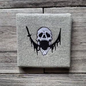 Puede incluir: Textil cuadrado gris con un diseño de calavera bordada. La calavera es blanca con detalles negros y una boca abierta. Dos espadas negras flanquean la calavera, con detalles negros goteando.