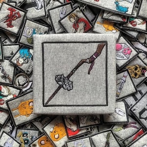 Peut inclure: Un écusson gris brodé avec un motif de bâton marron et violet avec une tête en pierre. Il est entouré d'autres écussons carrés avec divers motifs de personnages de dessins animés. Pour vêtements ou accessoires.