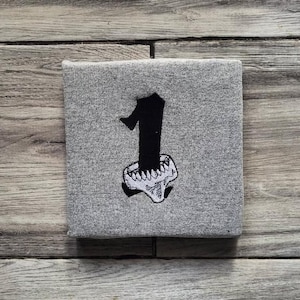 May include: A grey square fabric featuring a black number one design. The number one is in a gothic font style, embroidered above a skull-like design with teeth. The fabric is on a wooden surface.