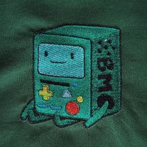 Könnte beinhalten: Gestickter Aufnäher des teal-farbenen BMO-Charakters aus Adventure Time, mit einem lächelnden Gesicht auf dem Bildschirm. Der Aufnäher befindet sich auf einem dunkelgrünen Stoff und zeigt ein gelbes Pluszeichen, einen roten Kreis und die Buchstaben "BMO".
