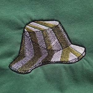 May include: Embroidered patch of a striped bucket hat in shades of olive green, grey, and white, with a black outline. The patch is sewn onto a green fabric background, creating a textured appearance.