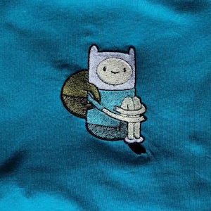 Peut inclure: Écusson brodé représentant Finn de la série Adventure Time. L'écusson montre Finn dans sa tenue emblématique, tenant un chapeau et un objet vert. L'écusson est cousu sur un tissu bleu sarcelle. Le design comprend du noir, du blanc, du sarcelle et de l'or.