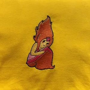 Puede incluir: Parche bordado con un personaje de dibujos animados con pelo naranja y un vestido rojo, sentado con los brazos alrededor de las rodillas. El parche está cosido con hilo naranja, rojo, rosa y amarillo sobre un fondo amarillo.