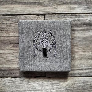 May include: A grey square textile featuring an embroidered skeletal design. The design includes a spiky halo, a rib cage, and a long, black, hair-like element. The textile is displayed on a wooden surface.