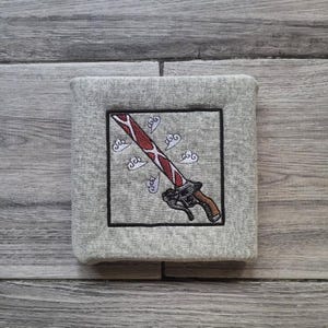 May include: A grey, square fabric item with an embroidered design. The design features a red and white sword with a black handle, set against a grey background with white cloud accents. The sword is framed by a black border.