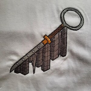 Può includere: Design ricamato di una spada con un'impugnatura circolare e una lama marrone e grigia. La spada ha una guardia dorata e una serie di piccoli fori lungo la lama. Il disegno è su tessuto bianco.