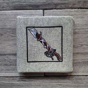 Könnte beinhalten: Ein graues, quadratisches Stoffstück mit schwarzem Rand, das ein gesticktes Design eines Schwertes mit braunem Griff, silberner Klinge und roten und weißen Akzenten zeigt. Der Artikel hat ein strukturiertes Aussehen.