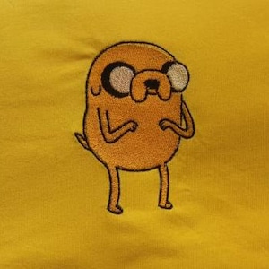 May include: Embroidered orange cartoon character on a yellow background. The character has large eyes, a small mouth, and a tail. The design is centered on the fabric.
