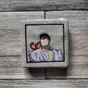 May include: A square gray fabric patch featuring an embroidered illustration of a character with spiky black hair, a red headband, and a light purple gi. The design is enclosed within a thick black embroidered square border on the gray fabric.