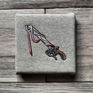 May include: Grey square fabric featuring an embroidered sword design. The sword has a brown handle, black details, and red and white accents, with a red ribbon attached. The fabric is on a wooden surface.