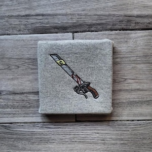 May include: A grey square textile featuring an embroidered sword design. The sword has a silver blade with a gold detail, a brown handle, and red accents. The textile is set on a wooden surface.