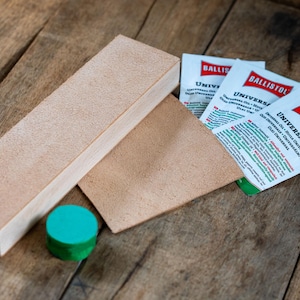 May include: A rectangular wooden block with a textured tan leather surface, a separate piece of tan leather, a small round green plastic cap, and three white sachets labelled Ballistol Universal Oil with red and black text and logos.